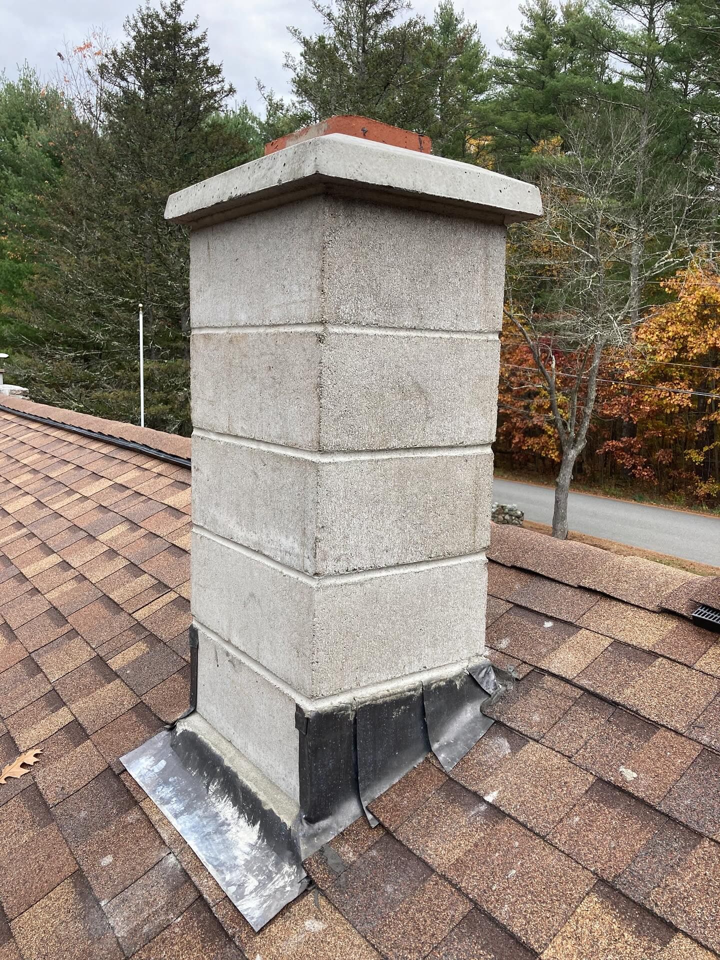 Gray block chimney on a brown shingled roof, with trees in the background.