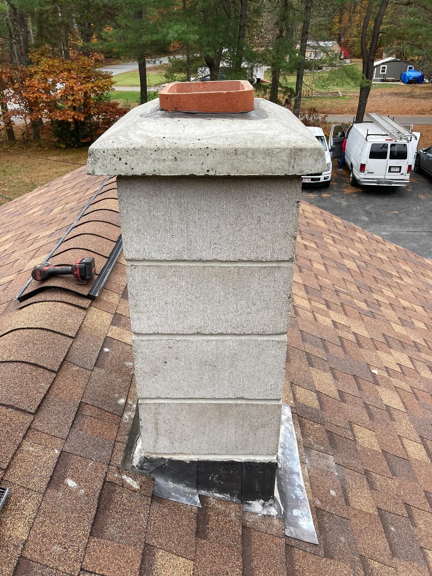 Chimney made of gray blocks on a brown shingled roof, topped with a concrete cap and red clay flue.