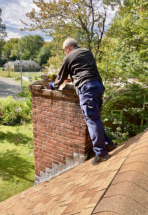 Person on a rooftop, working on a brick chimney; blue pants, black jacket, and gray hair.
