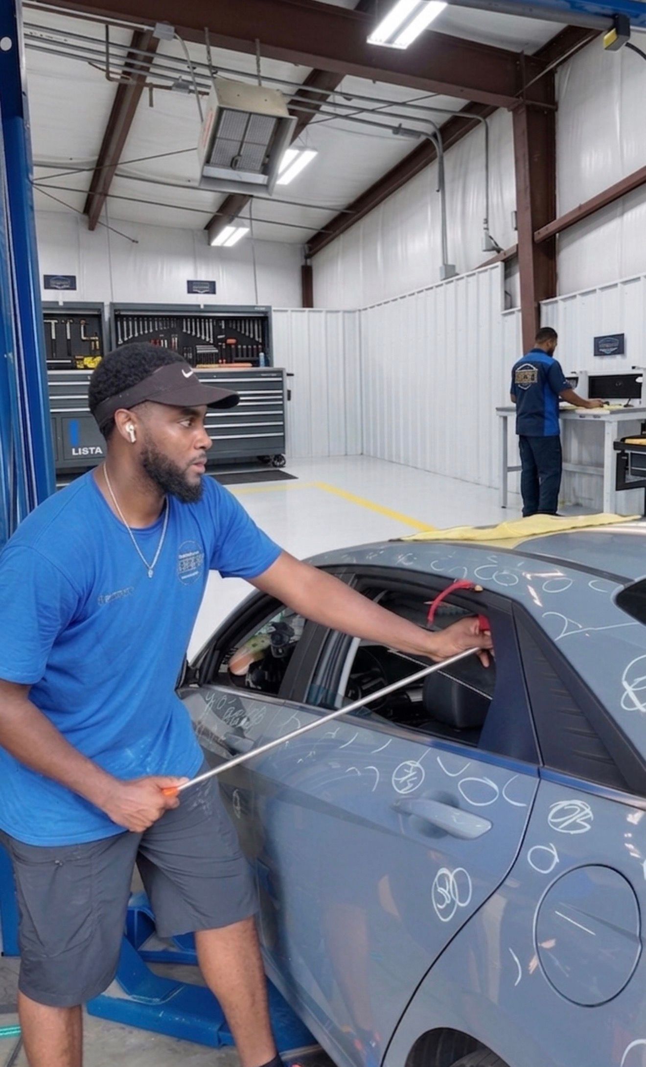 A technician in a blue Charleston dent company shirt and visor holds a long PDR tool while working on the roof area of a grey car covered in white circles (circles indicate where the  dents are). 
