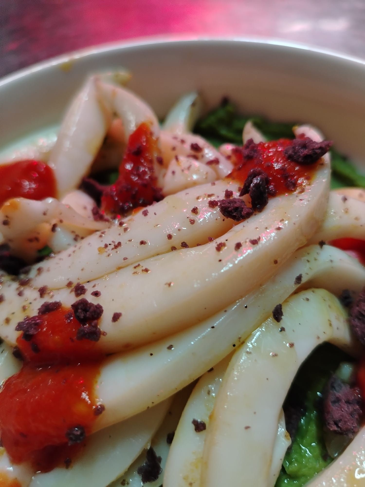 Primo piano di un piatto di calamari bianchi con salsa rossa e granella scura. Si intravedono delle foglie verdi.