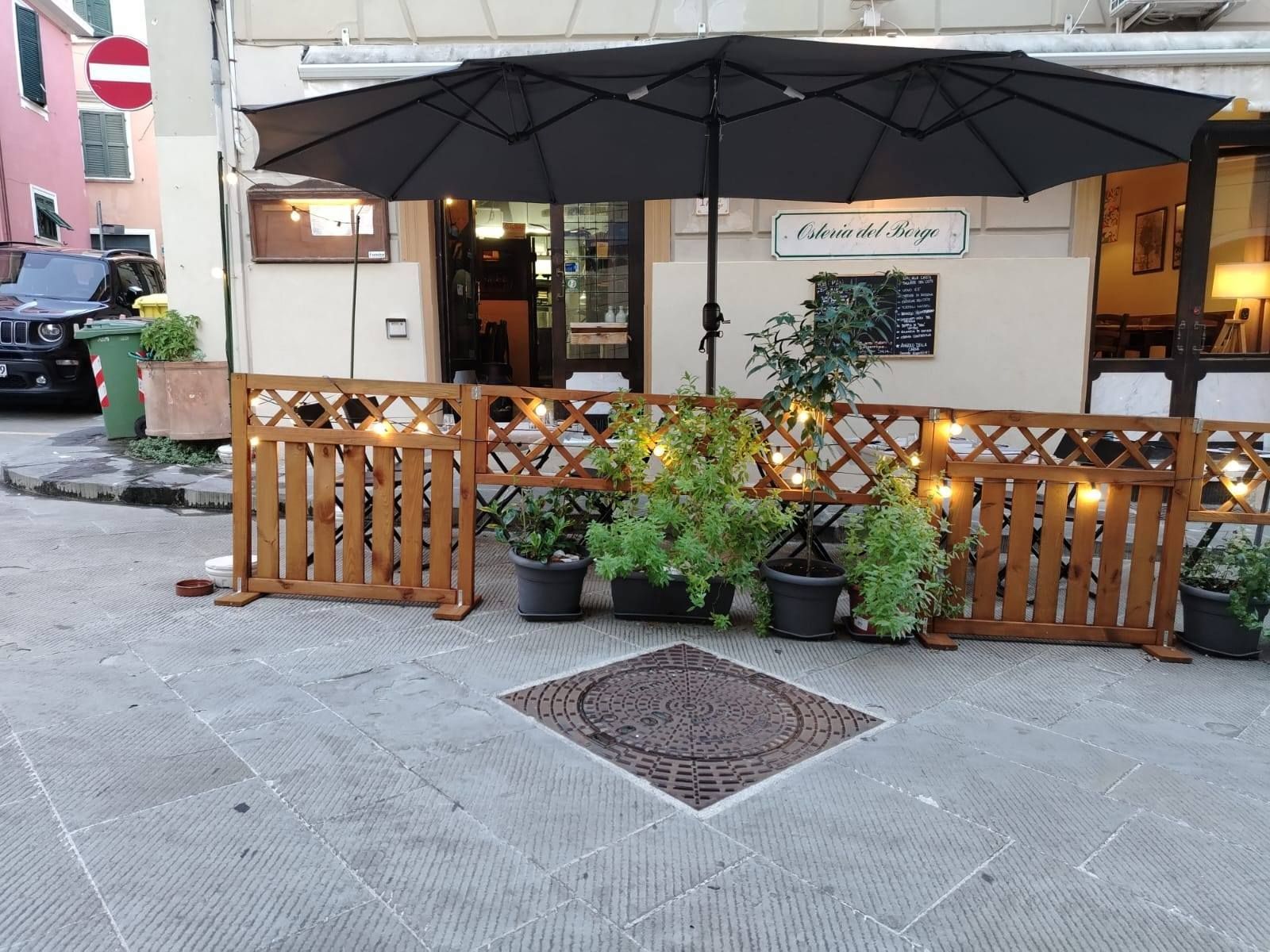 Posti a sedere all'aperto del ristorante con staccionata in legno, ombrellone nero, piante e luci a stringa.
