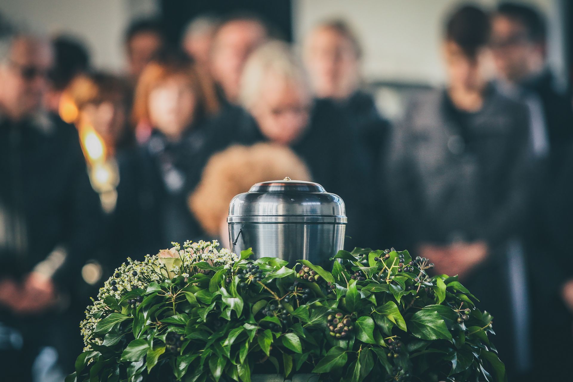 Metal urn surrounded by green foliage at a memorial service, crowd blurred behind