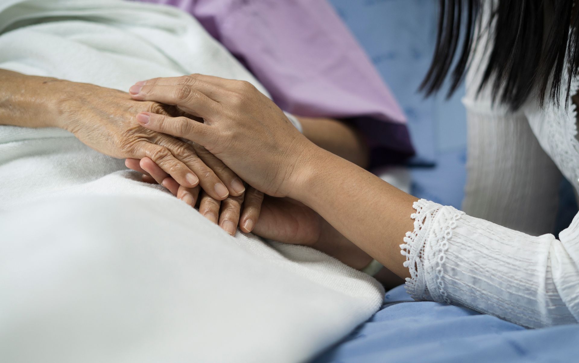 A person's hand clasped by another, both on a hospital bed.