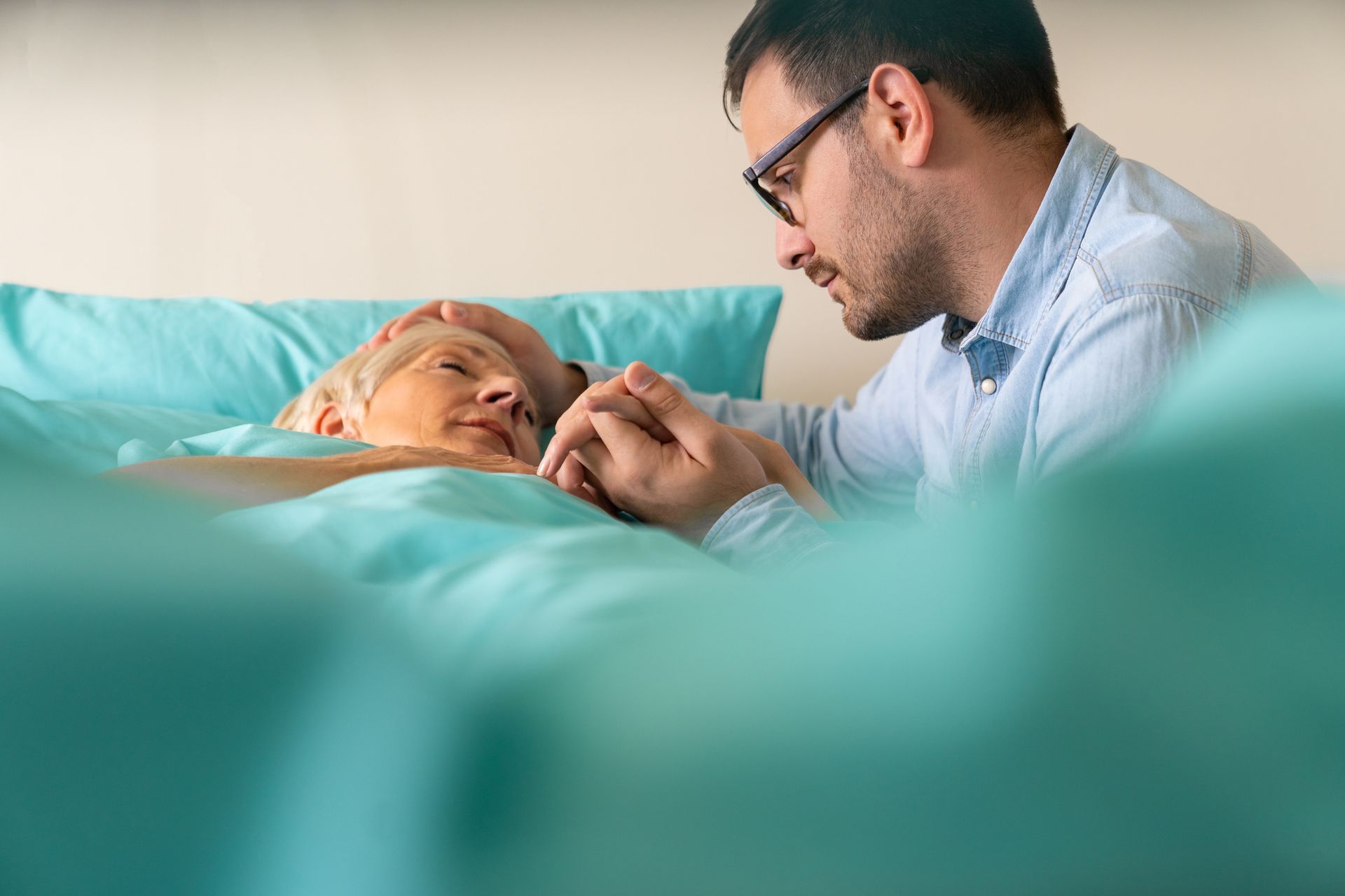 Man holding hands with person in bed; soft lighting, teal bedding.