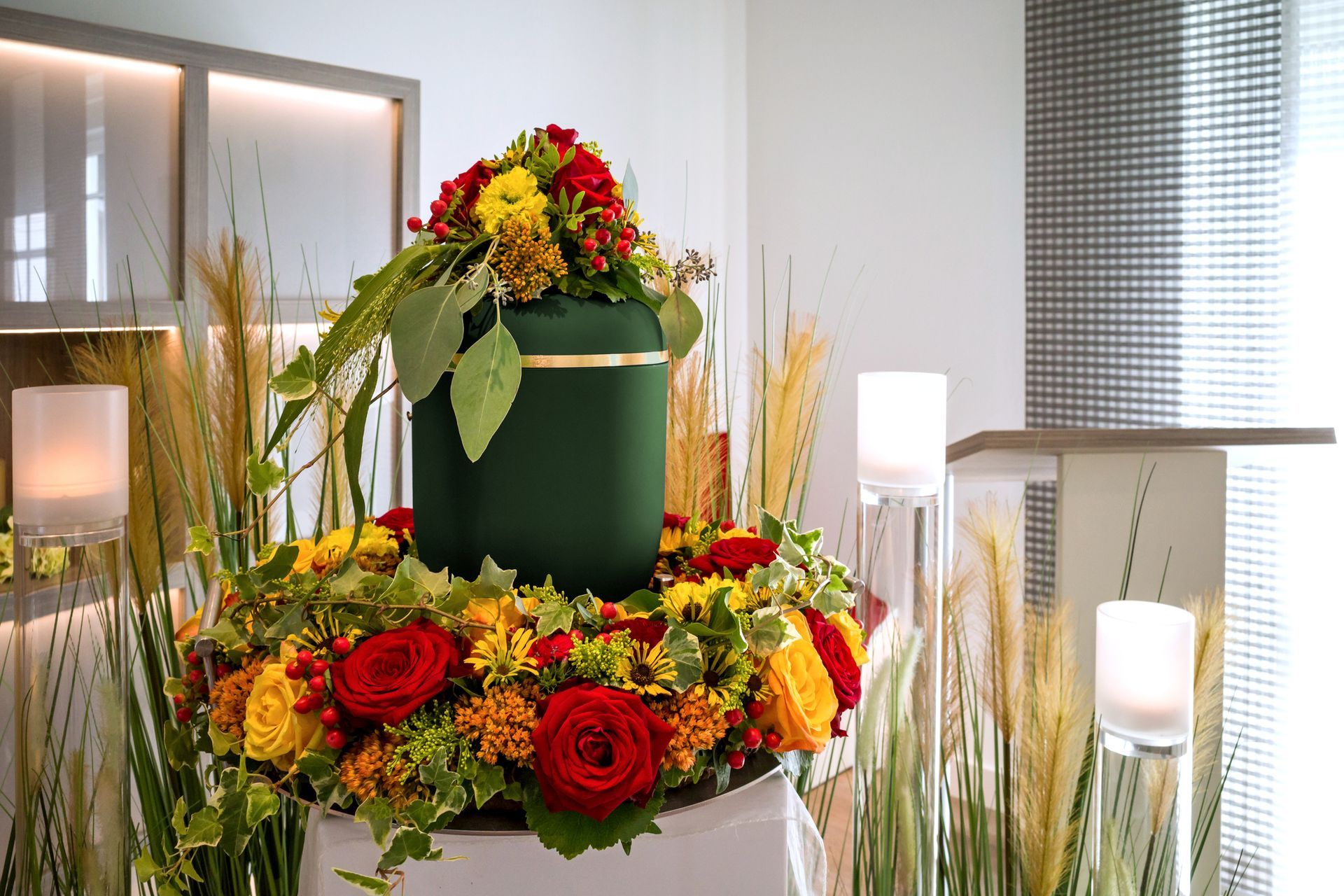Green urn with floral arrangement, surrounded by flowers, candles, and tall grasses.