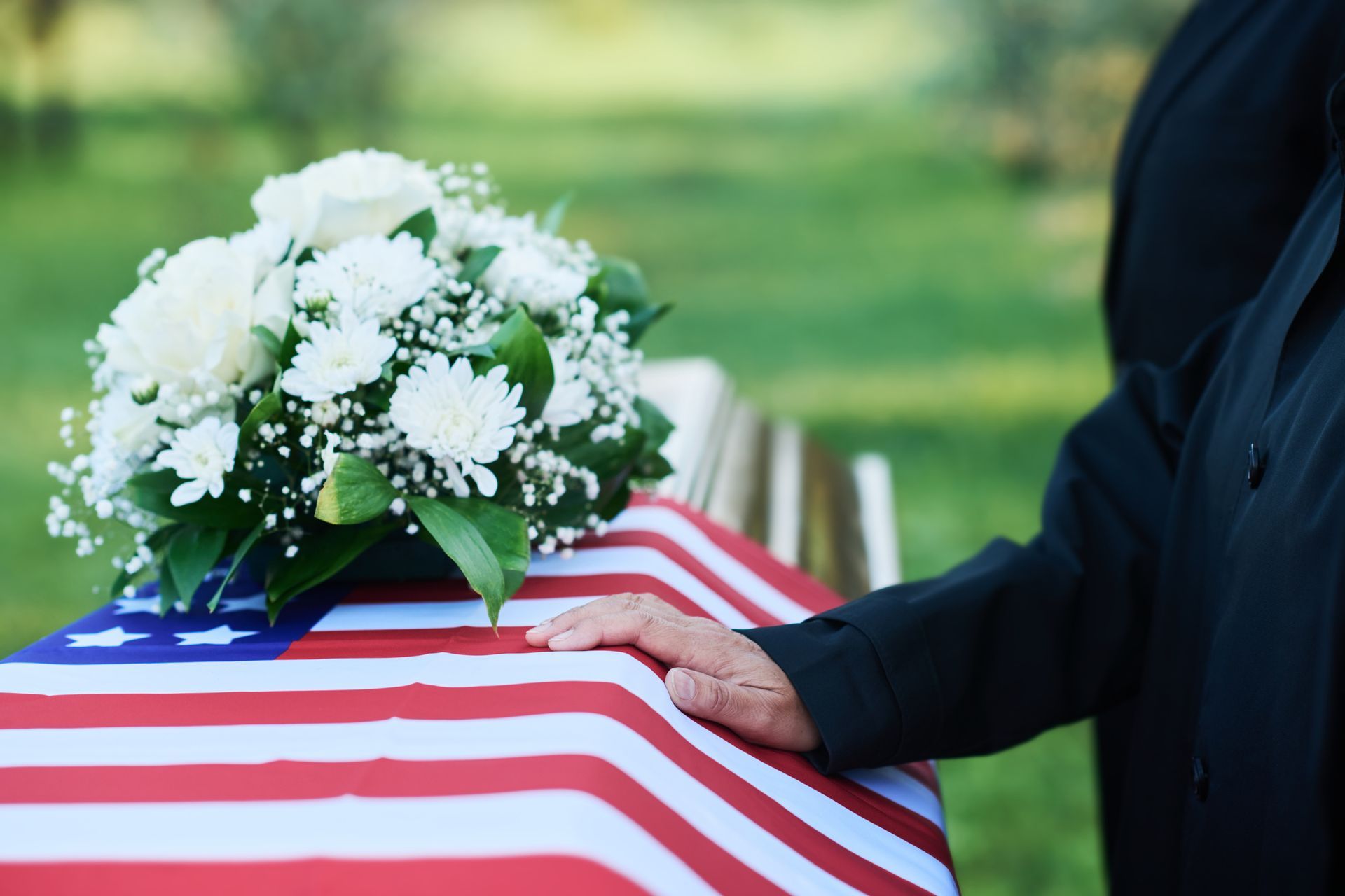 Coffin draped with American flag, topped with flowers, person's hand resting on it. Outdoor setting.