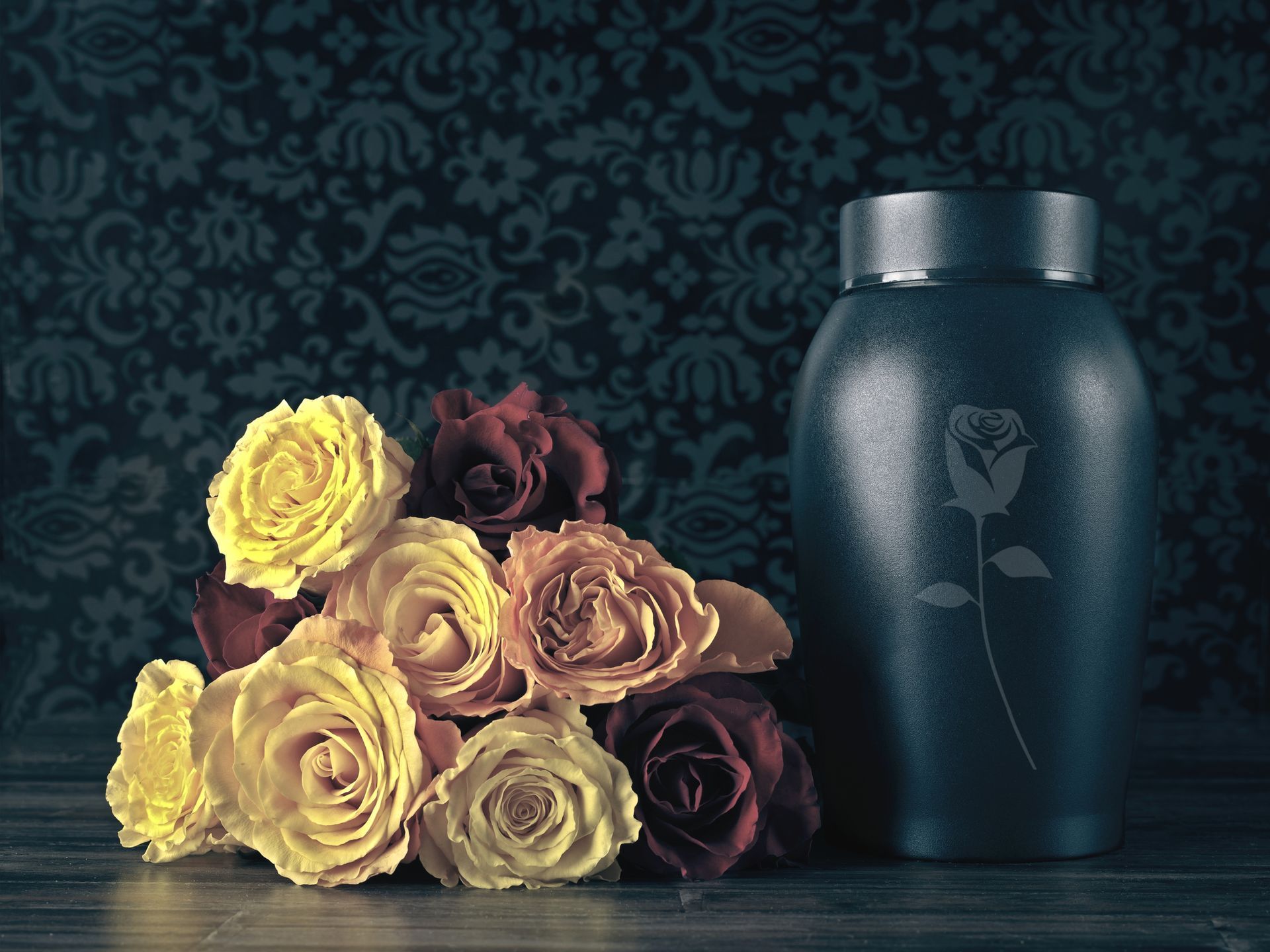 A bouquet of yellow, orange, and red roses next to a dark blue urn with a rose etched on it.