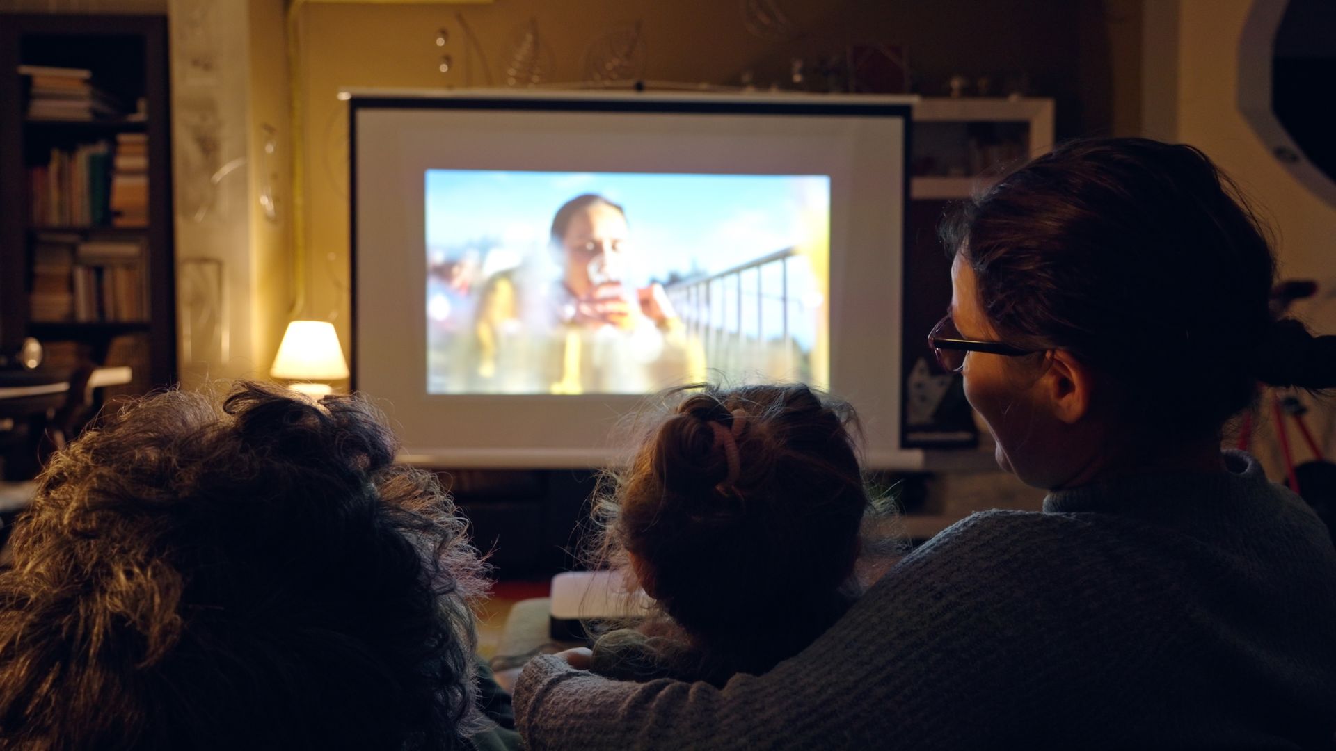 Three people watching a movie projected on a screen in a dimly lit room.