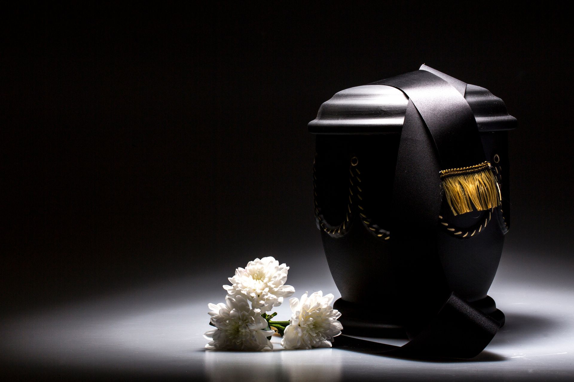 Black cremation urn with ribbon and flowers, lit by a spotlight, on a dark background.