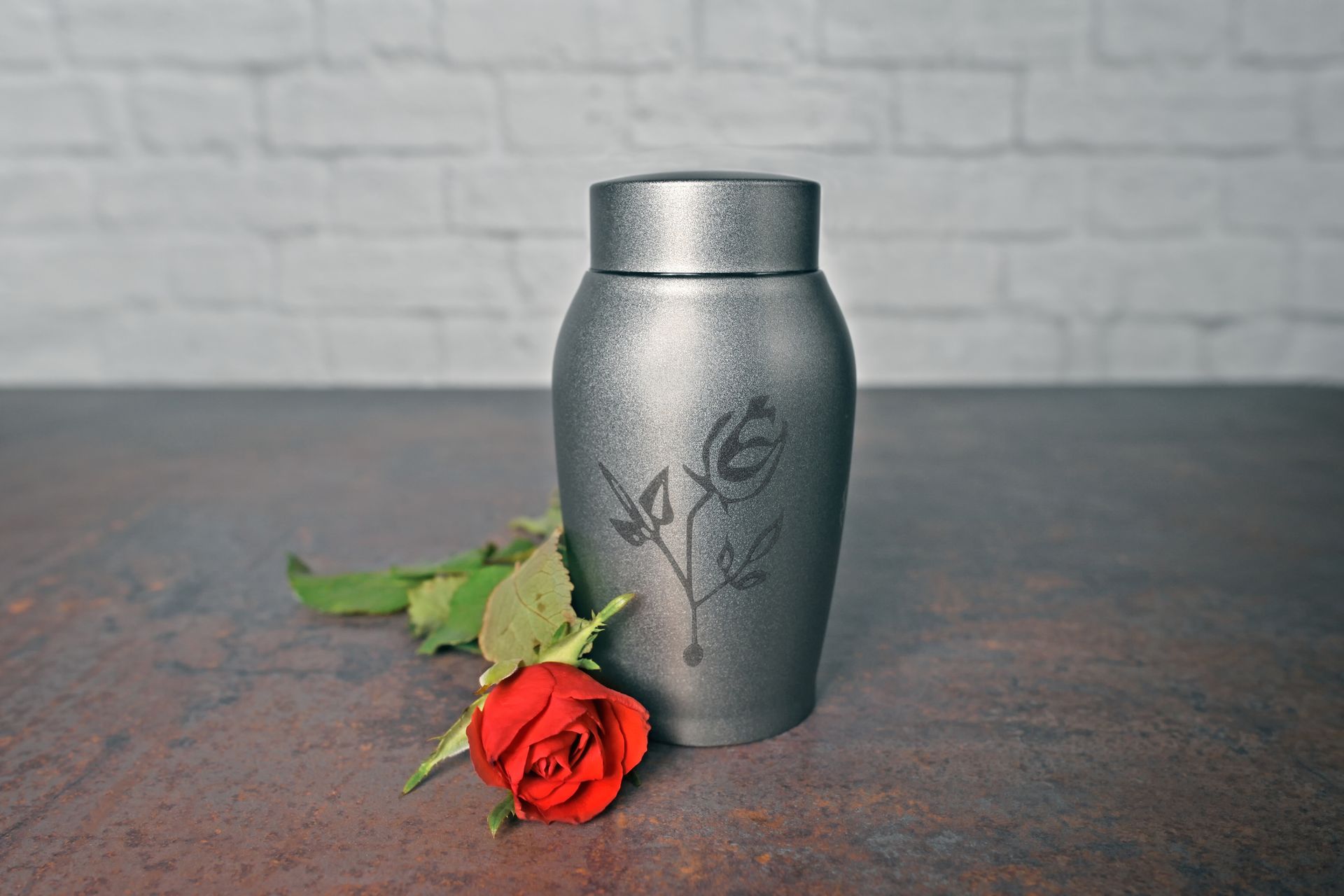 Silver urn with etched rose design, red rose and leaves. On a mottled, brown surface with a white brick backdrop.