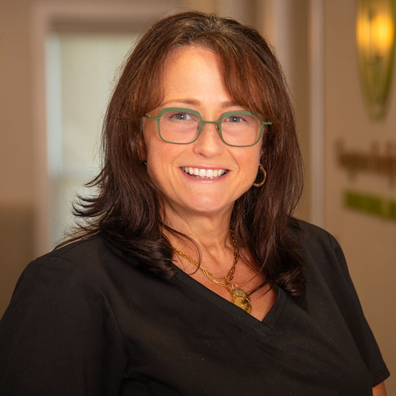 Woman with glasses smiles, wearing black shirt and gold jewelry. Indoors, blurred background.