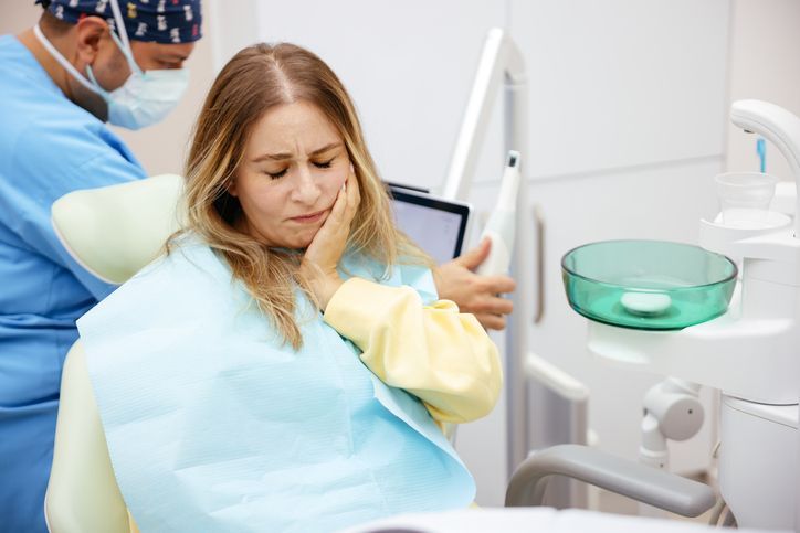 Woman in dental chair, holding cheek in pain; dentist in scrubs attends.
