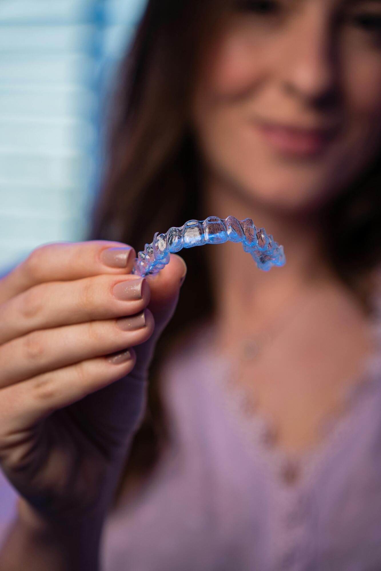 Hand holding a clear blue dental aligner in front of a blurred person indoors