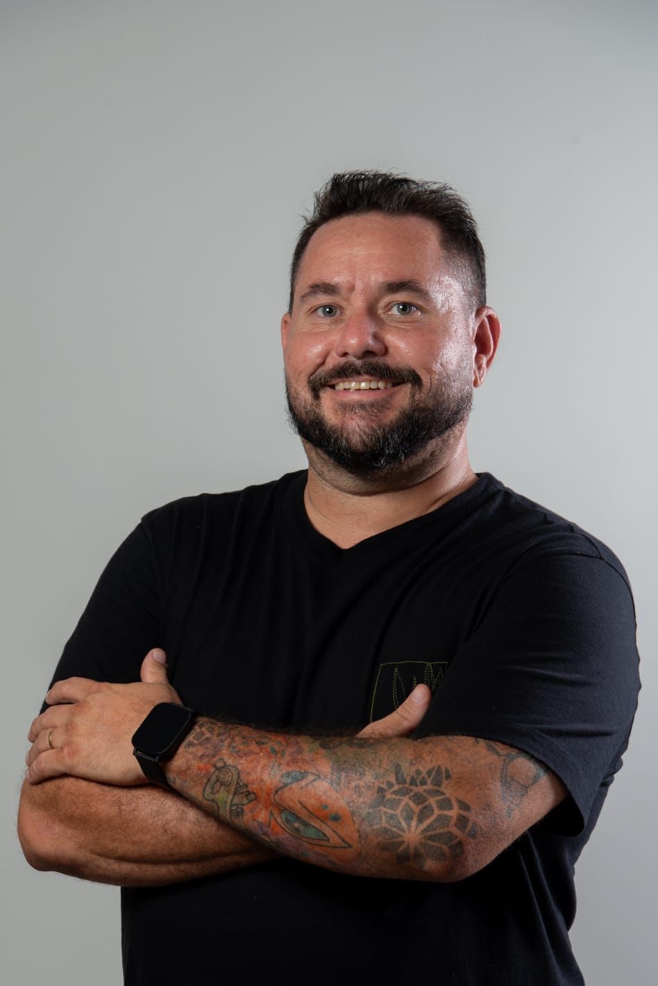 Homem com tatuagens sorri, braços cruzados, vestindo uma camisa preta, contra um fundo cinza.