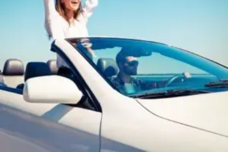 Woman with arms raised in a white convertible car on a sunny day. Driver at the wheel.