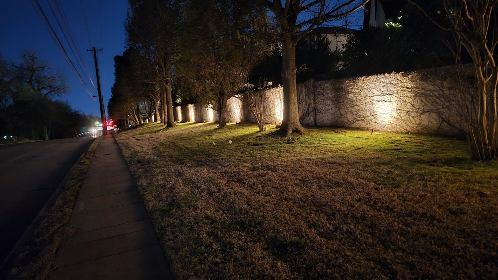 A stone wall illuminated by warm landscape lighting at night, bordered by a sidewalk and trees beside a road.
