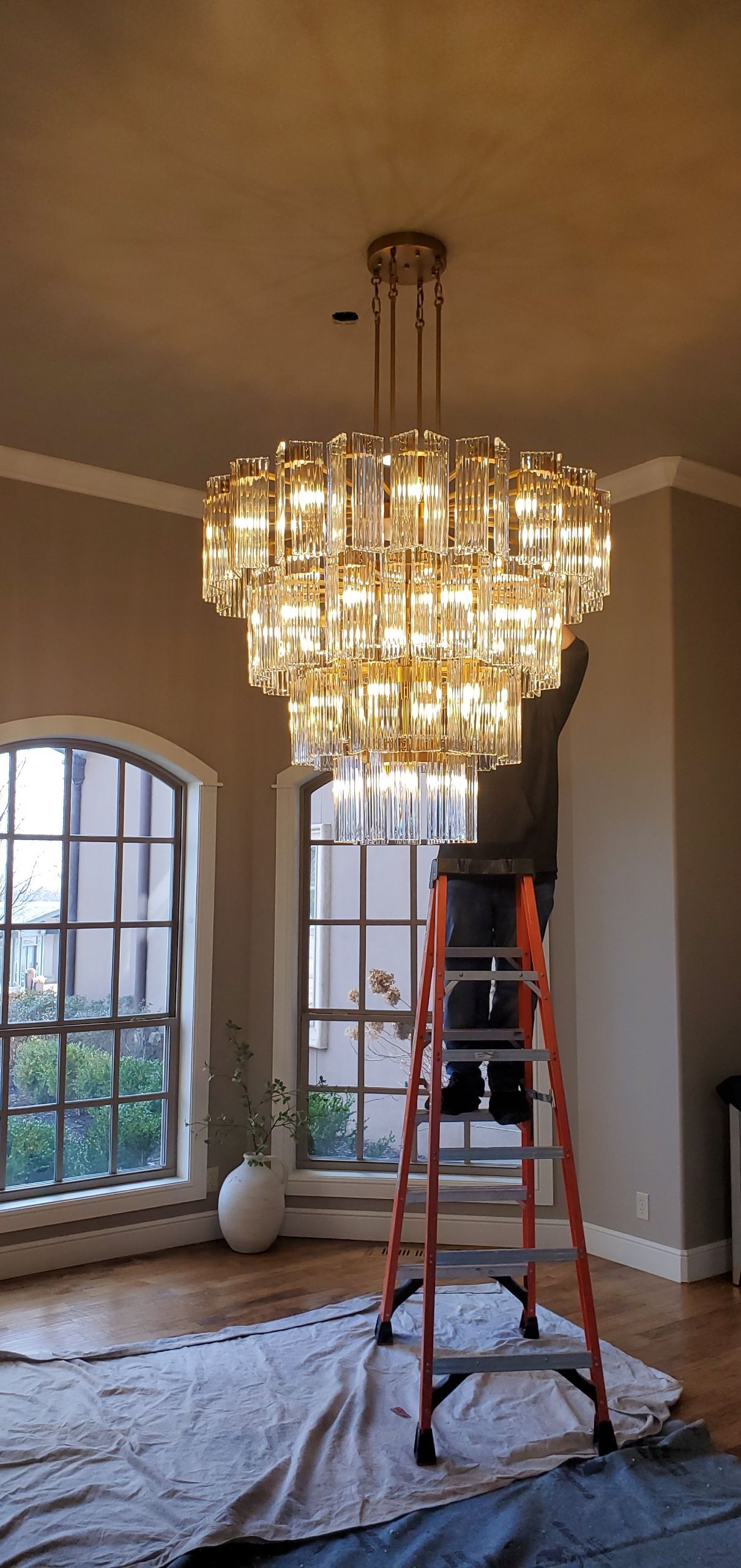 A multi-tiered crystal chandelier hangs in a room where a person on a ladder is installing or cleaning it.