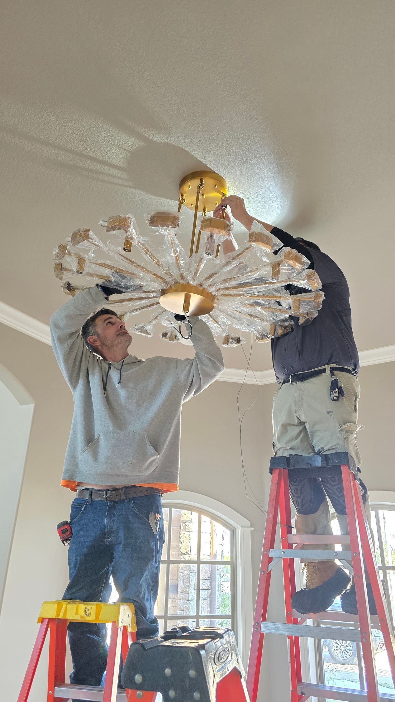 Two people on ladders install a large, round crystal chandelier onto a ceiling in a room with a window.