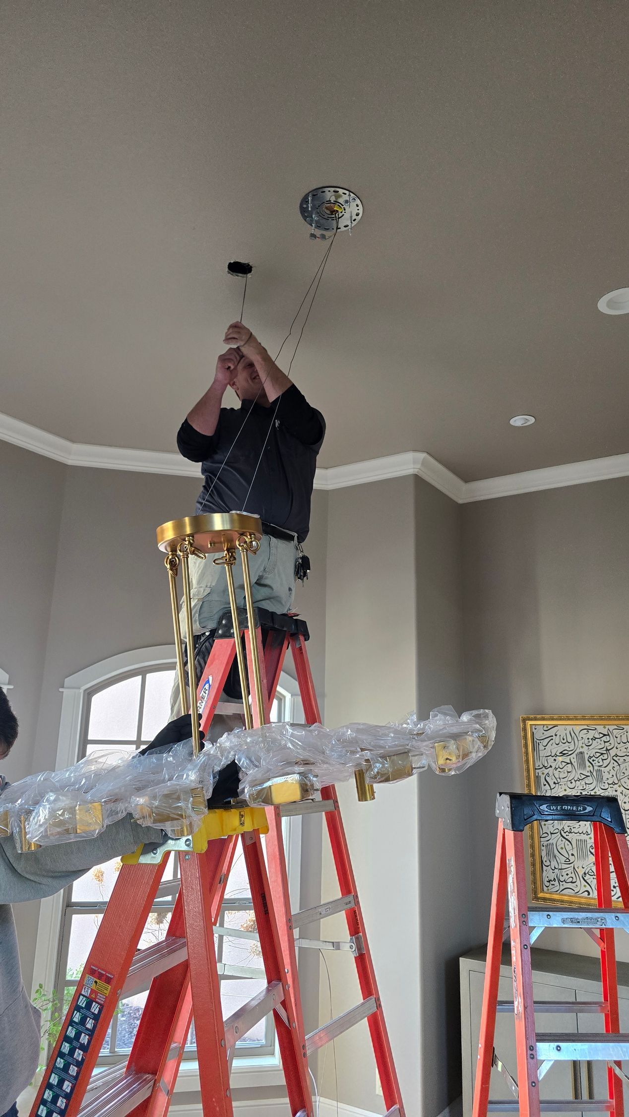A person stands on a tall ladder, installing a gold-colored chandelier into a ceiling electrical box in a room.