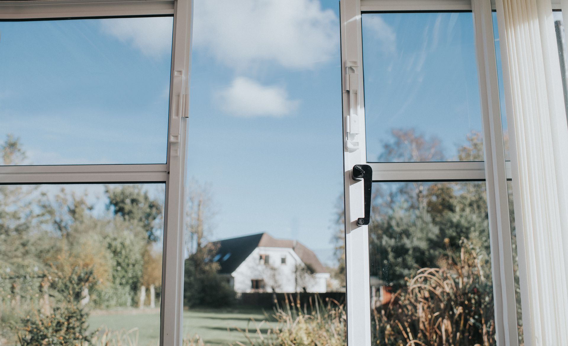 View through a window of a bright blue sky with a few clouds, a house, and trees. White window frame.