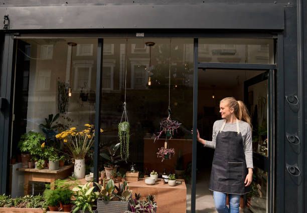 Woman in apron opens door to a flower shop filled with plants and arrangements.