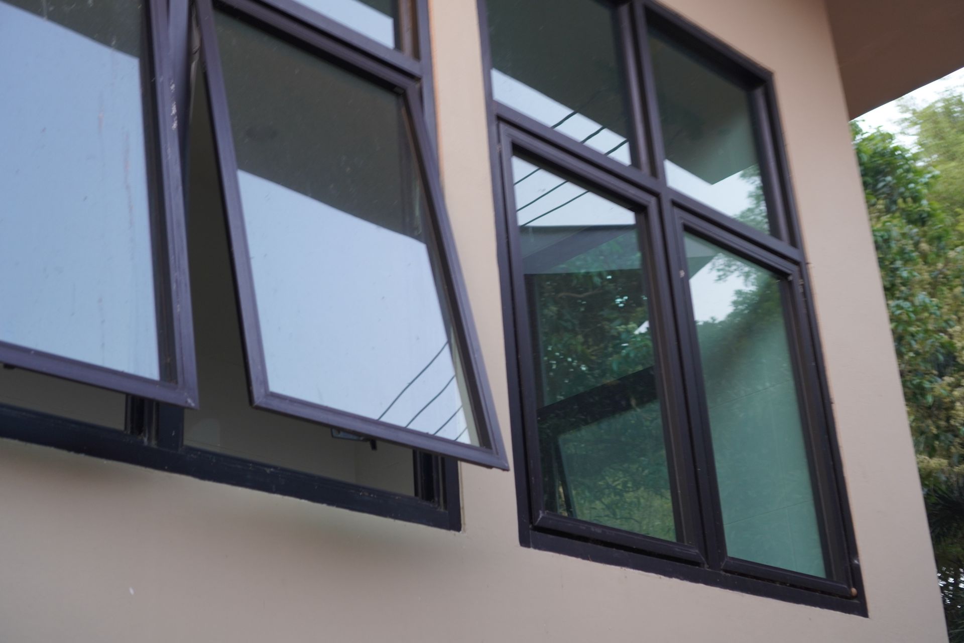 A close-up of a building's exterior shows black-framed windows, one partially open, against a beige wall, with trees visible in the reflection.