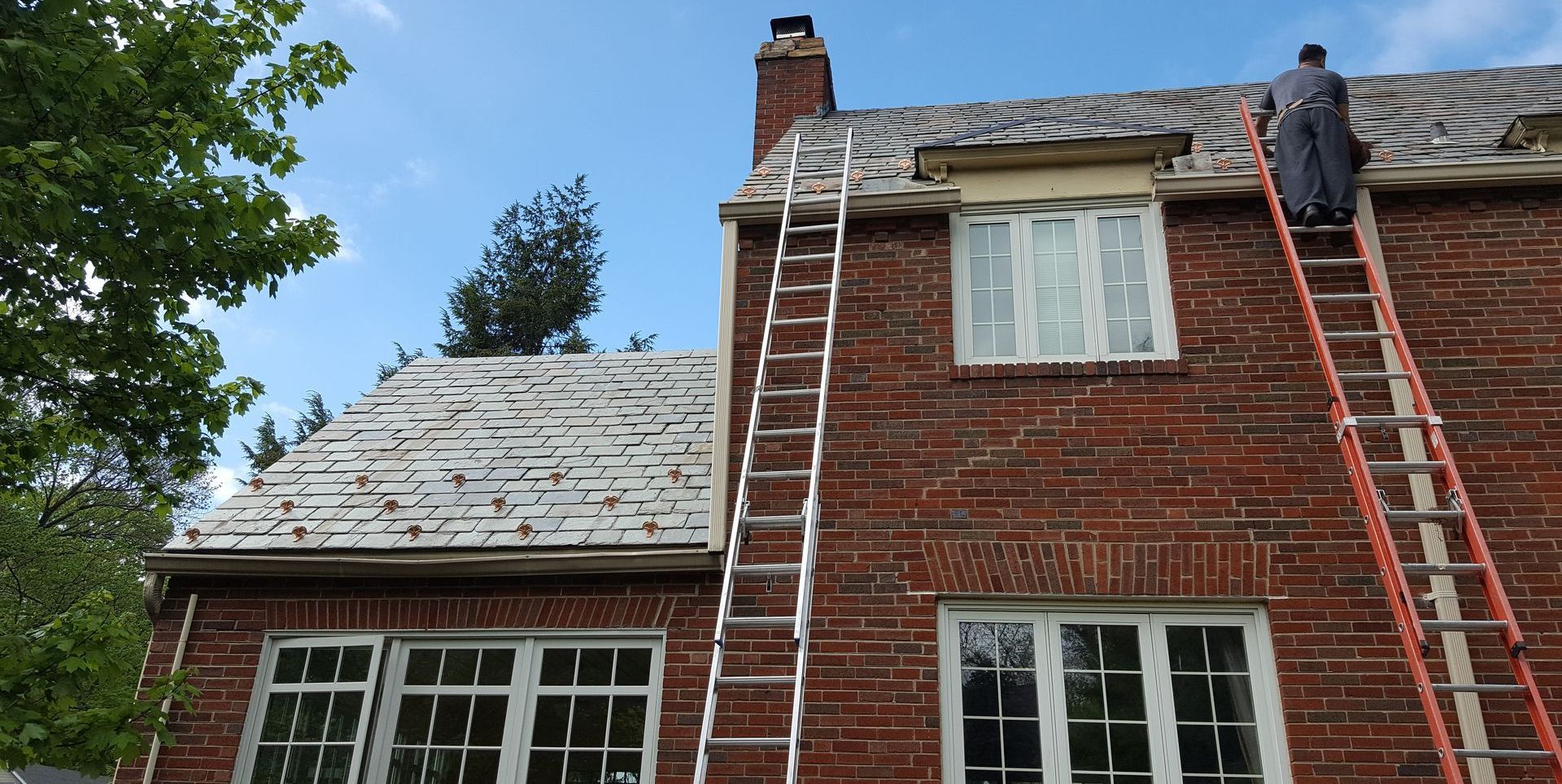 A man on a ladder is working on the roof of a brick house.