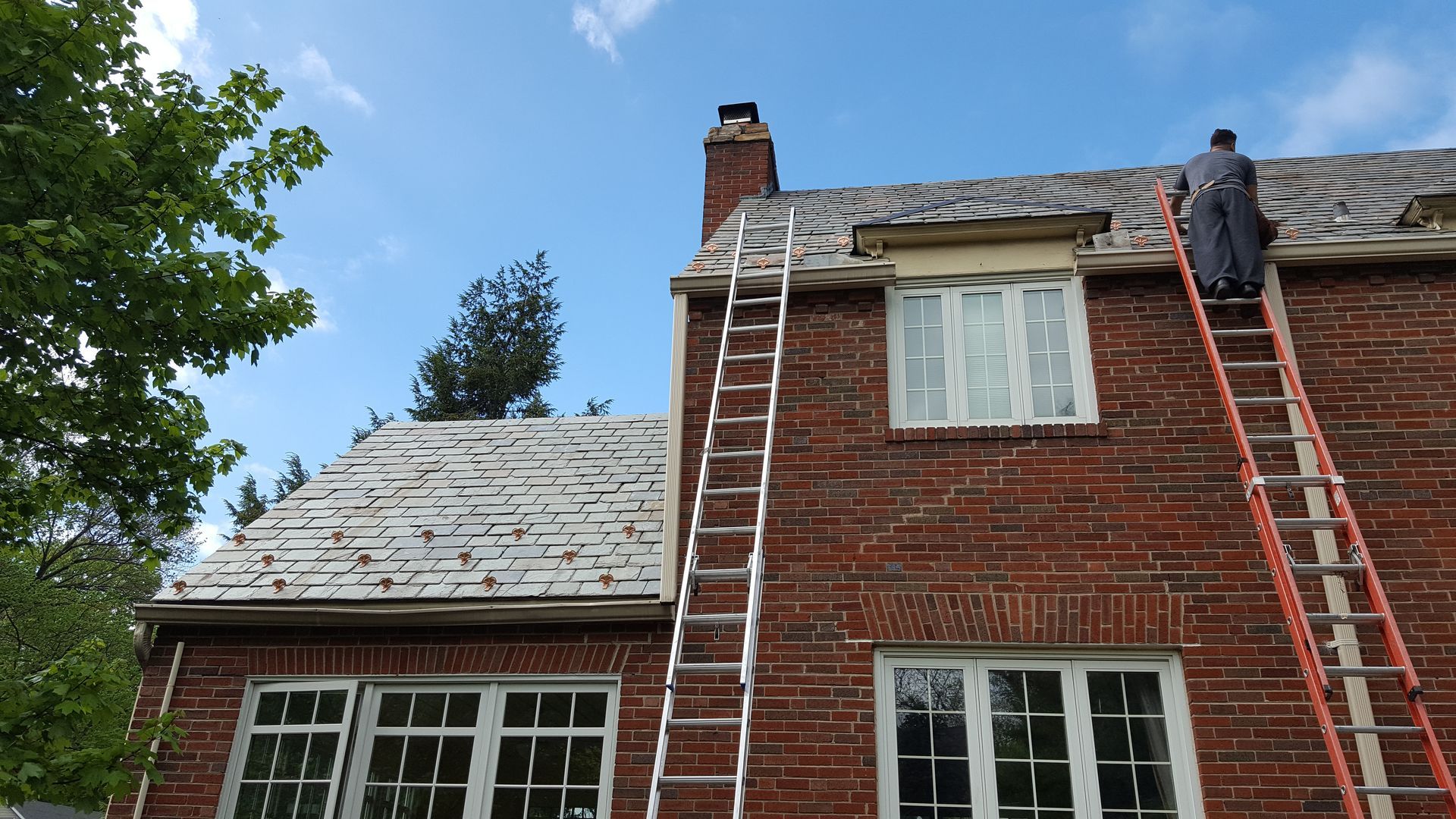 A man is standing on a ladder on the roof of a brick house.