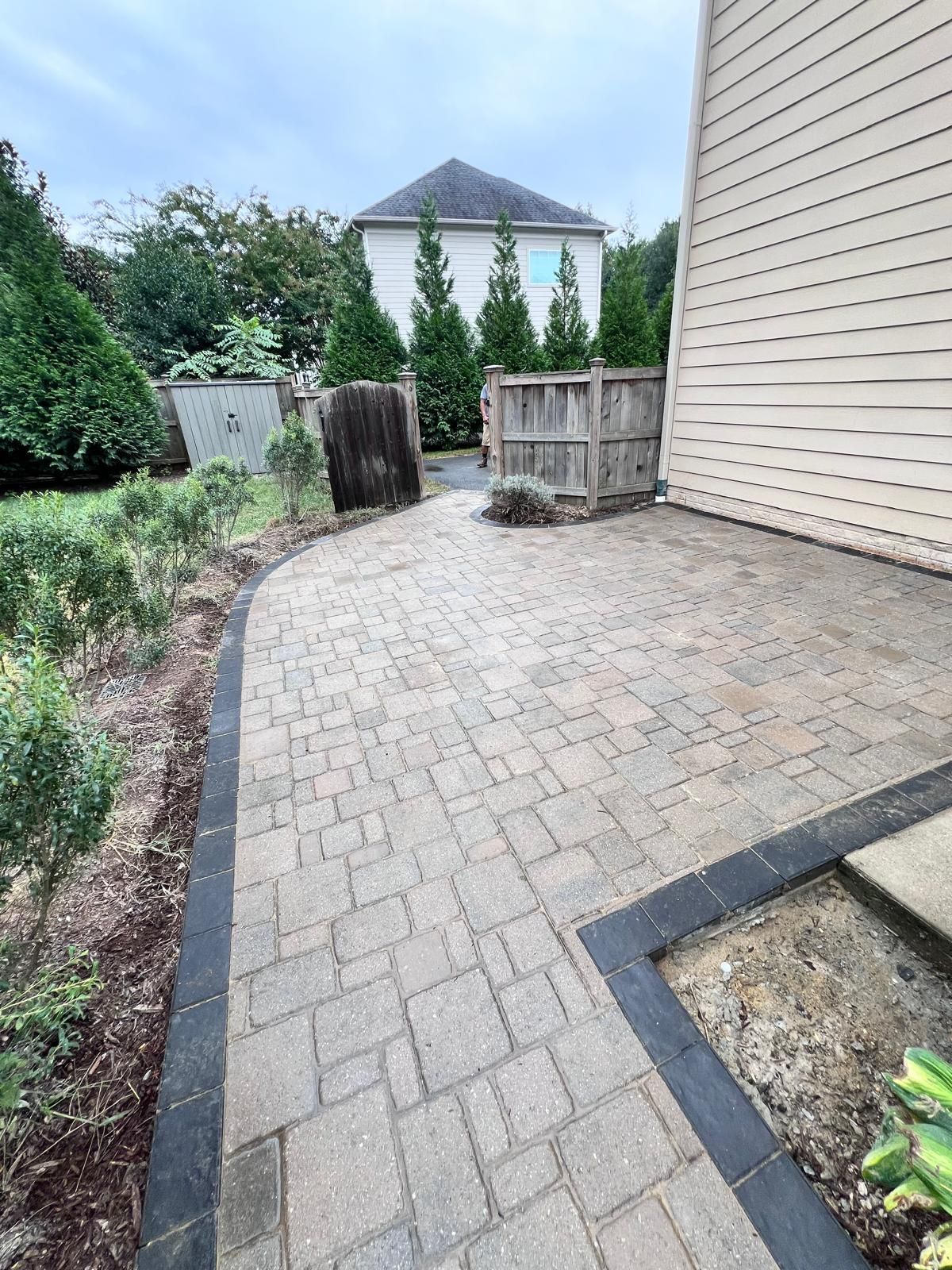 Brick patio with dark border, leading toward wooden structures near a beige house.