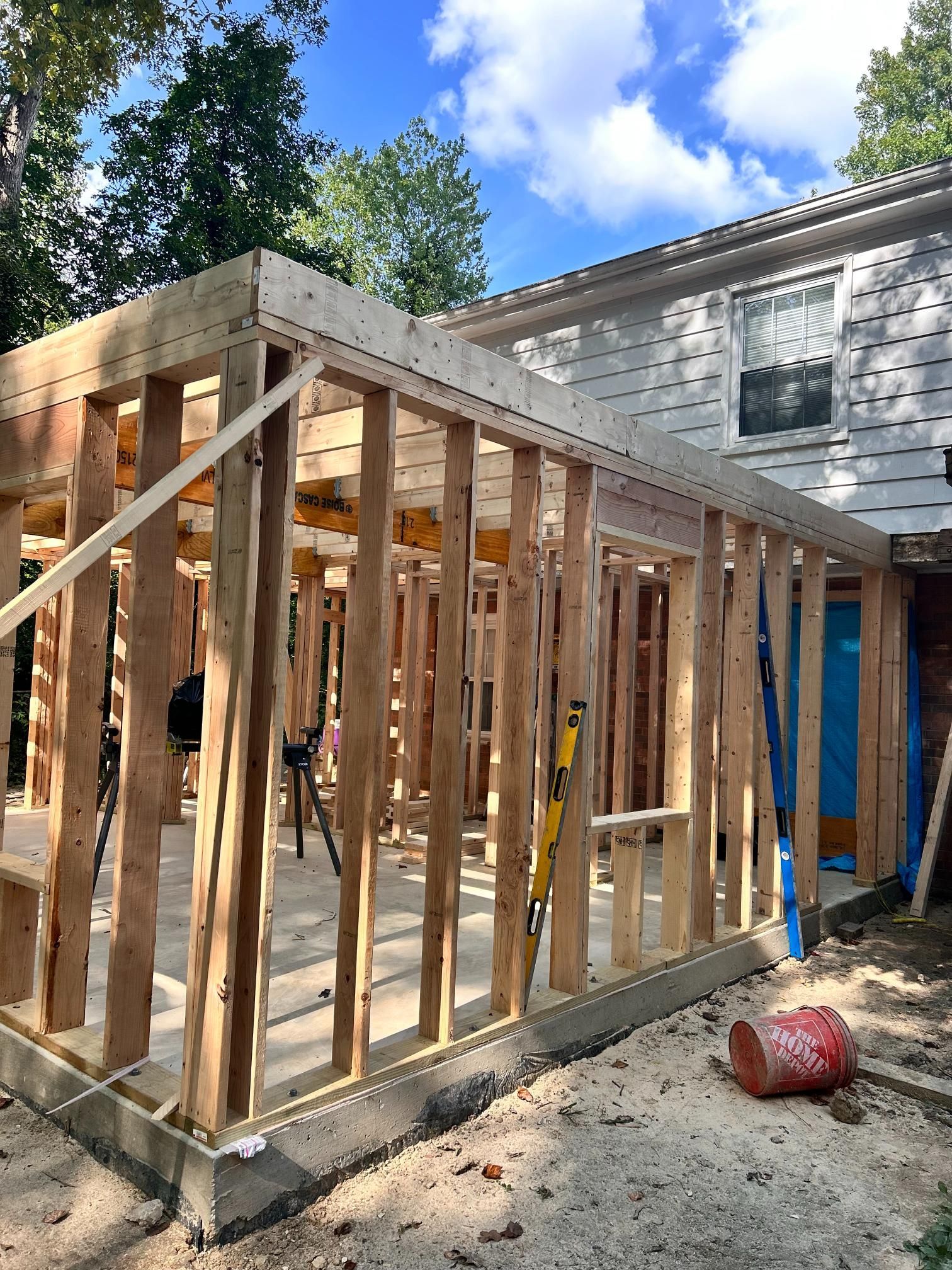 Wooden framing of a building addition under construction attached to a house, on a concrete foundation, with a red bucket nearby.