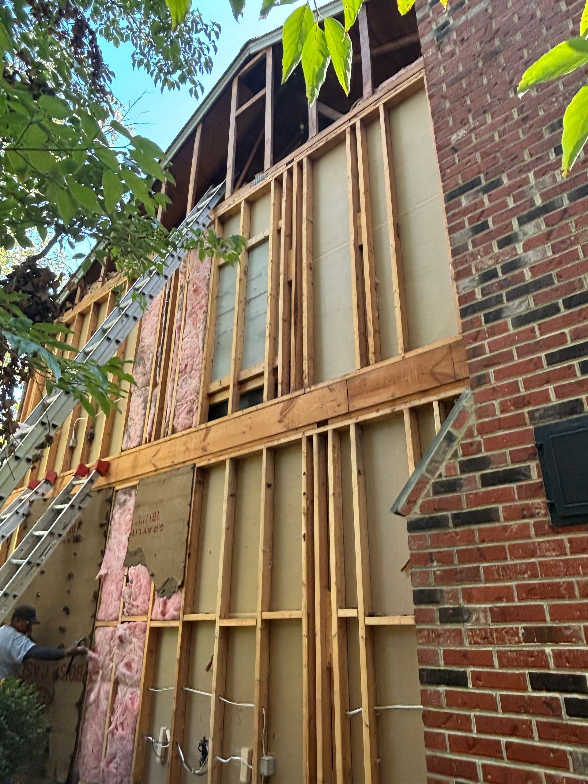 Construction on a two-story house. Exterior wall framing, insulation exposed, adjacent brick facade. Man working on side.