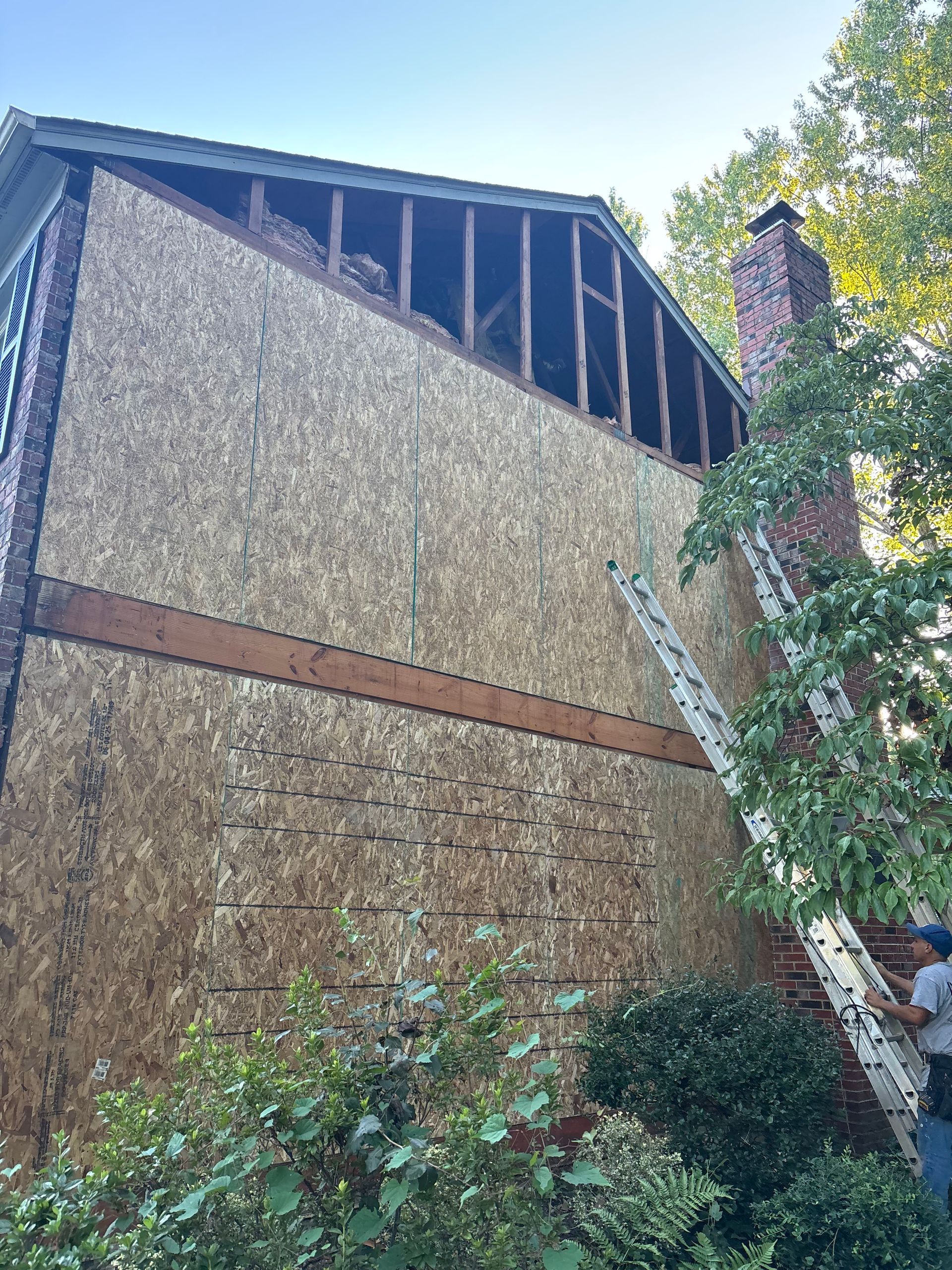 Side of house with exposed framing and OSB sheathing; ladder near a brick chimney.