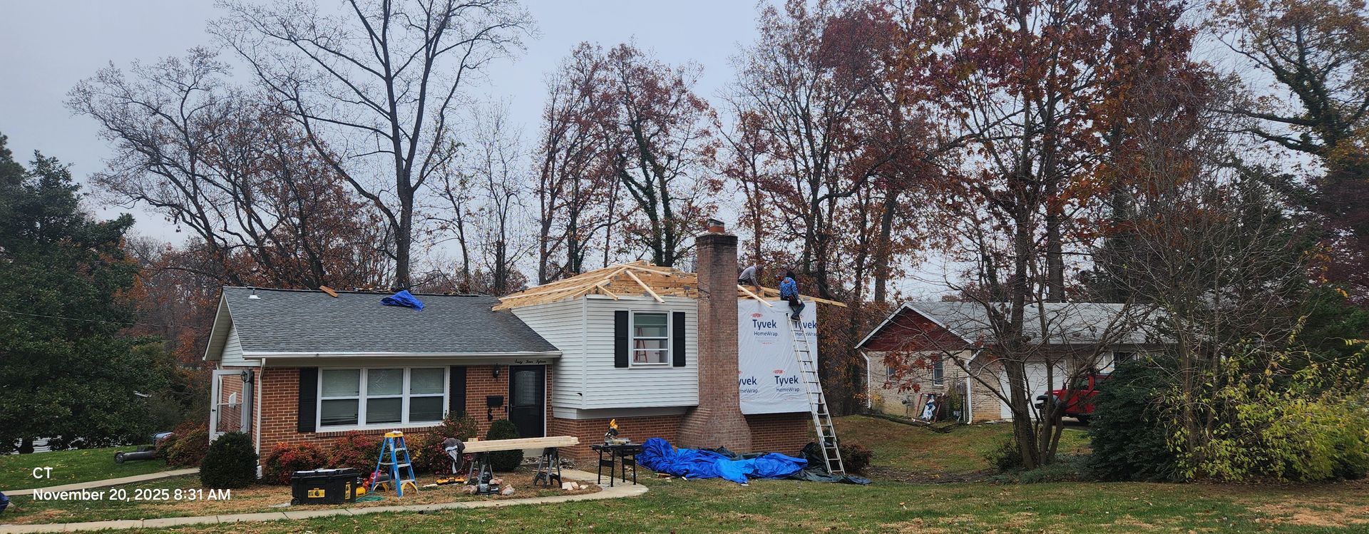 House under construction; workers on the roof. Brick and white siding with exposed roof beams, surrounded by trees.
