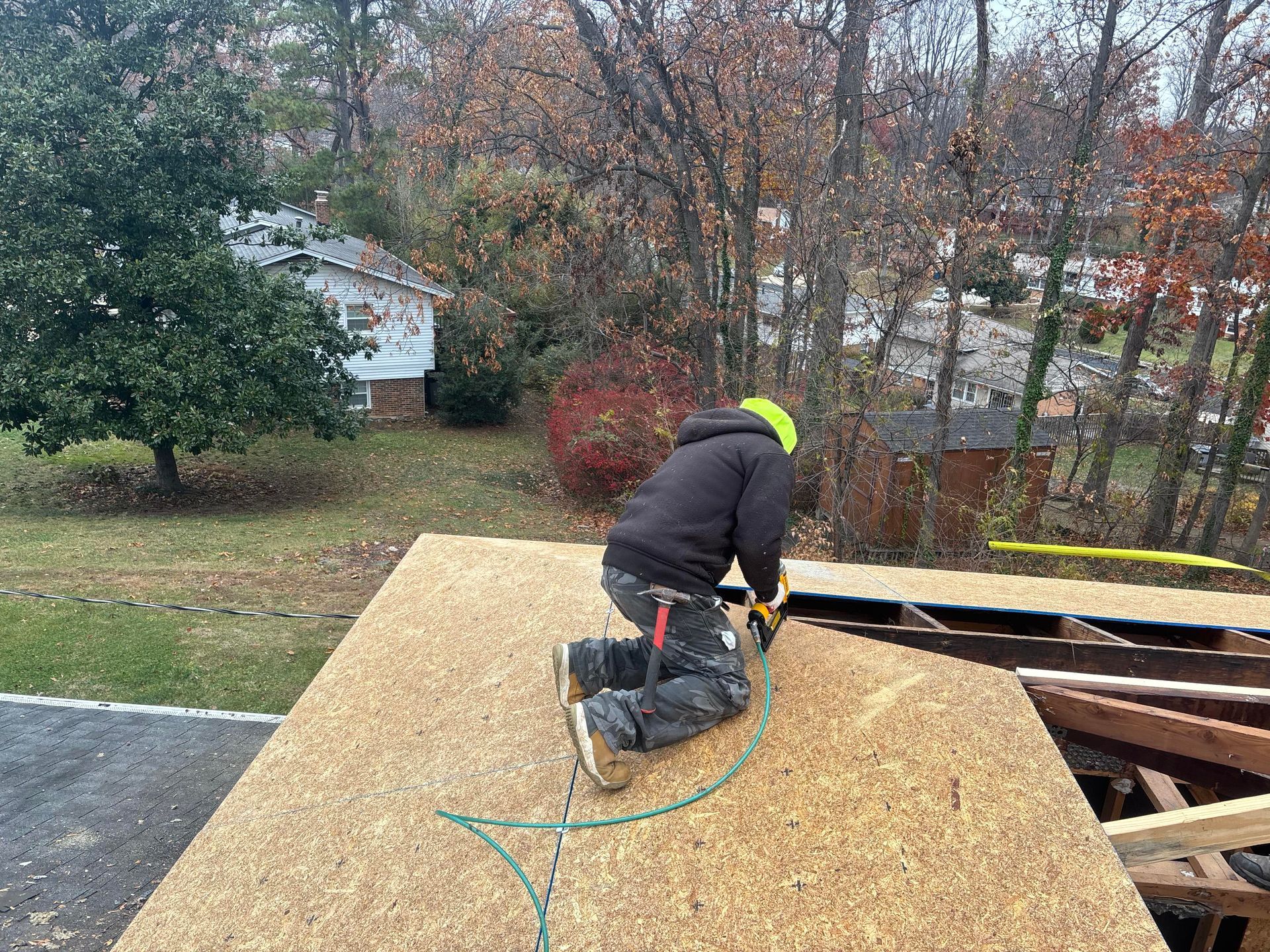 Roofer kneeling, sawing plywood on a roof; wearing a black jacket and hard hat. Trees and houses in background.
