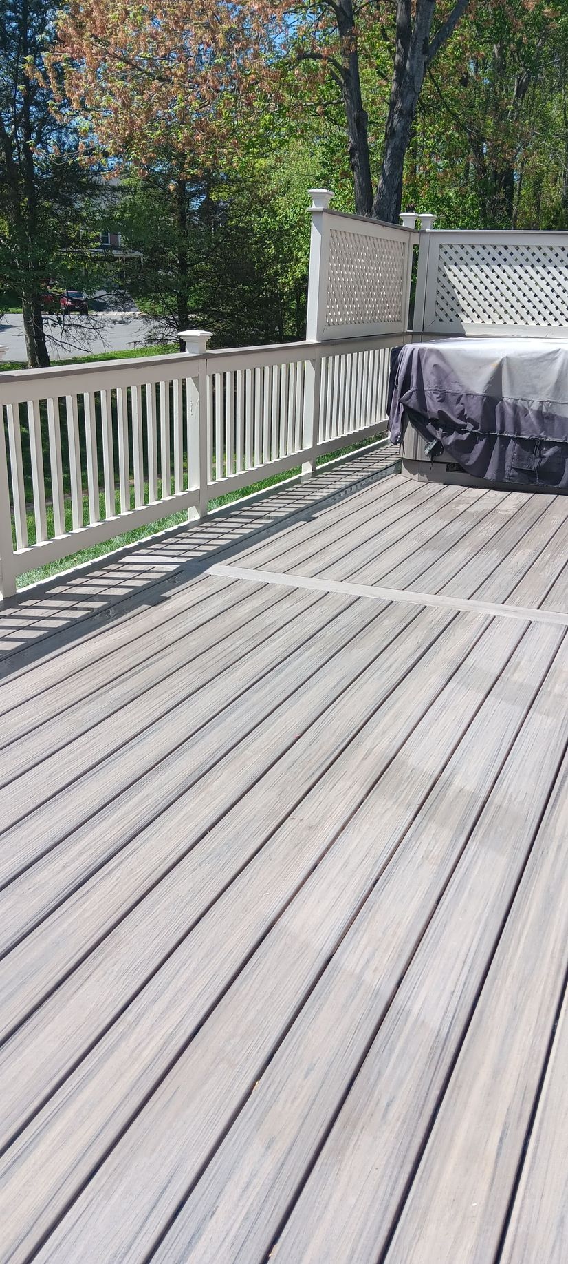 Wooden deck with white railings, trees, and a covered hot tub. Sunny day.