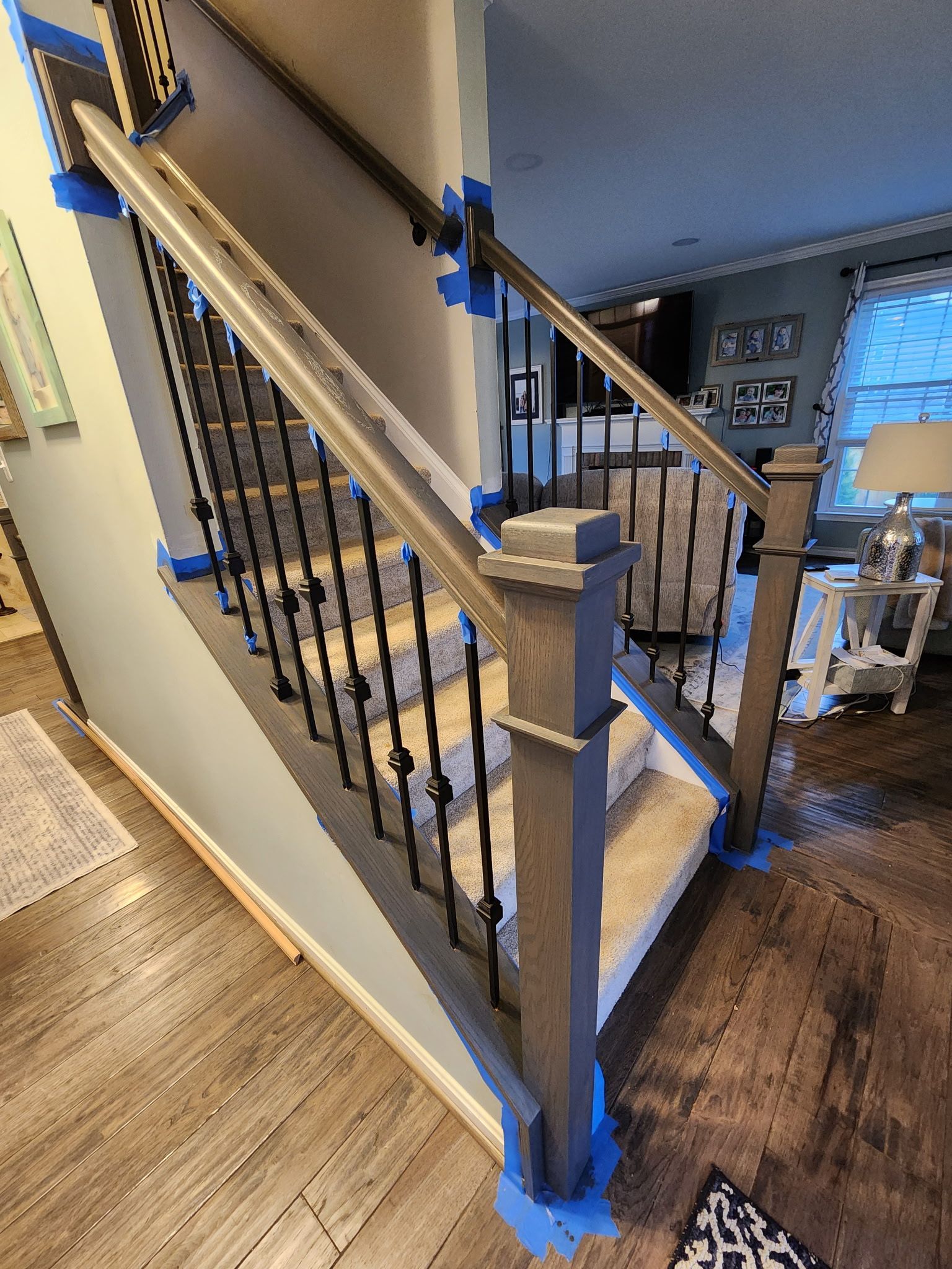 Staircase with gray wooden handrails and black metal spindles. Blue tape protects adjacent surfaces during a painting project.