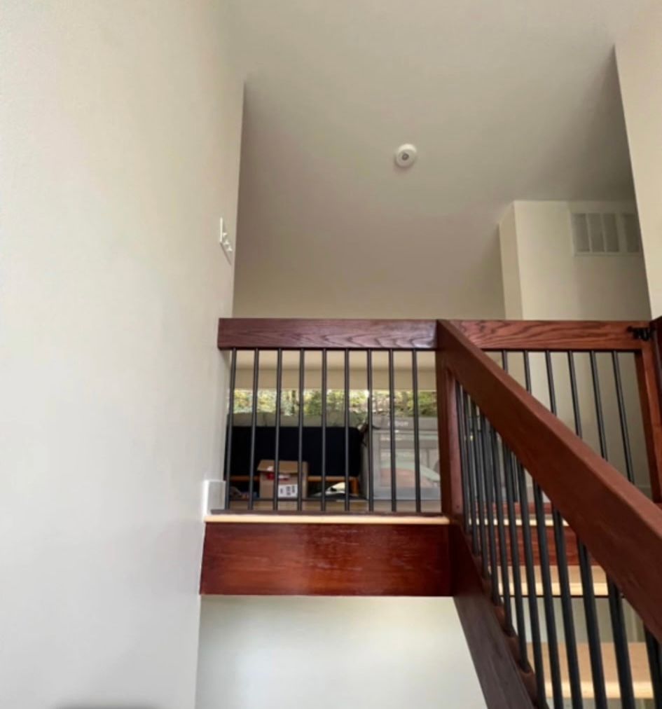 Interior view of a staircase with dark wooden railing and metal bars. A smoke detector is on the ceiling above.