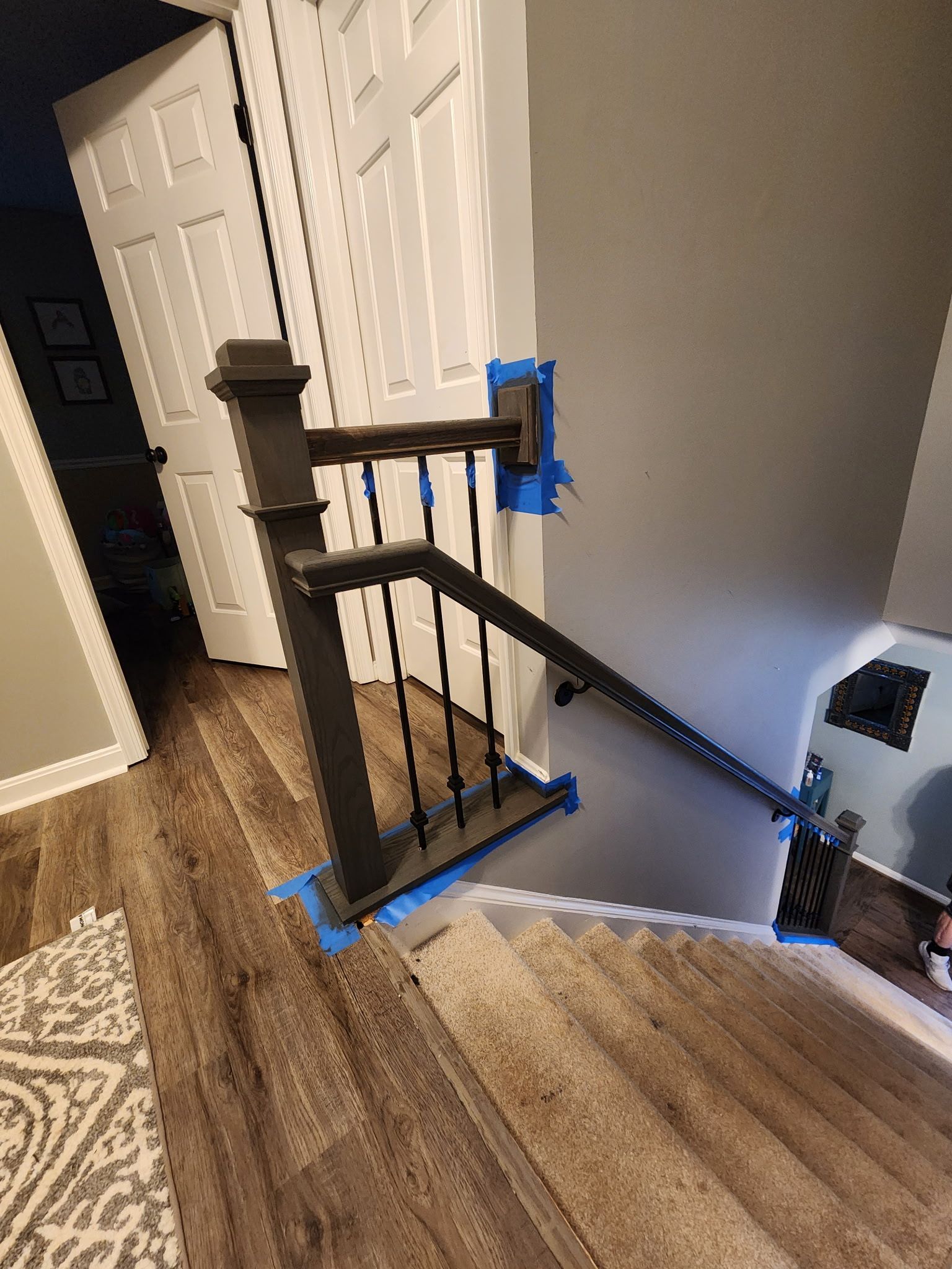 Staircase with dark spindles and handrail, blue tape for painting. Wooden floors and gray walls.