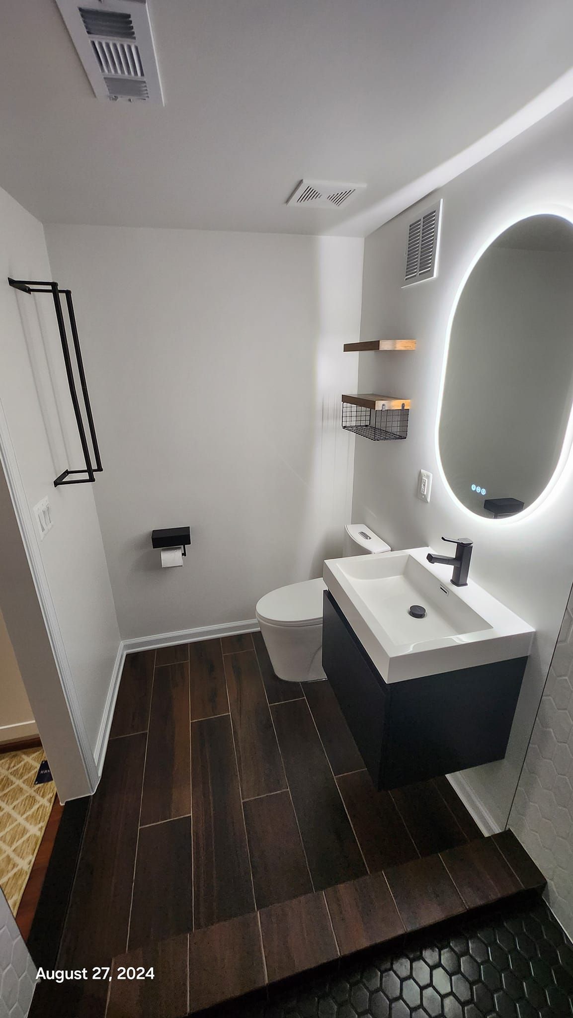 Small, modern bathroom with dark wood floors, white walls, black vanity, and an oval mirror.