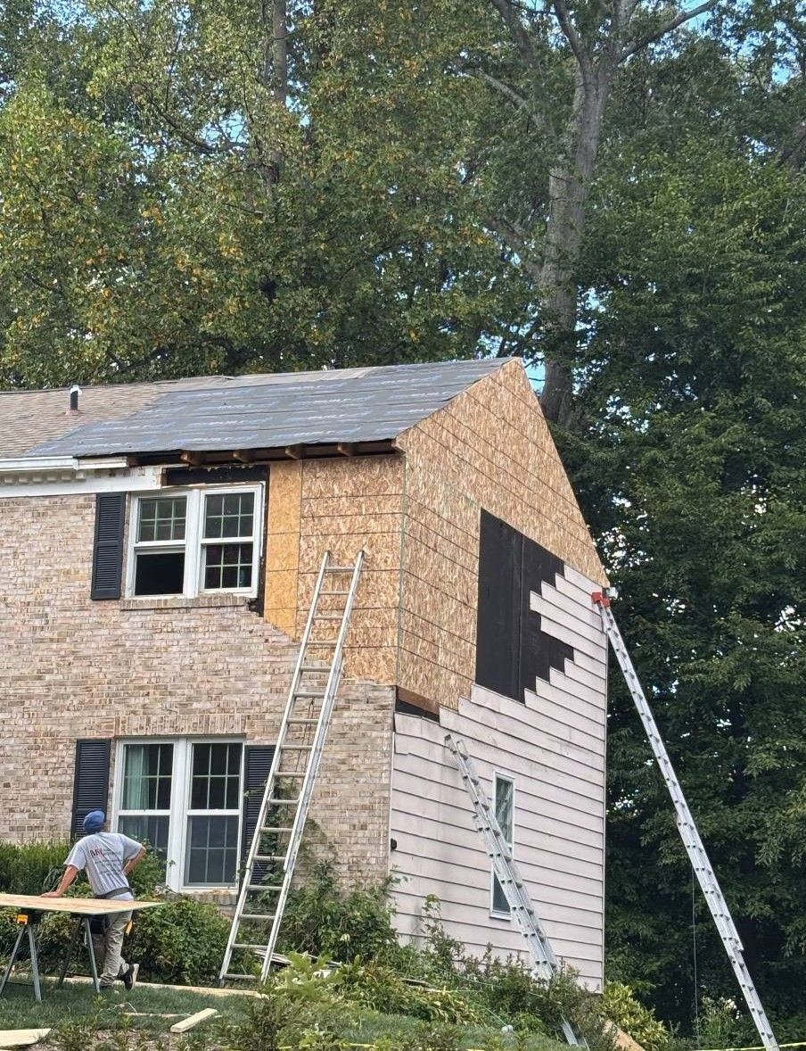 Construction workers siding a two-story brick house. Ladders, exposed wood, and trees are visible.