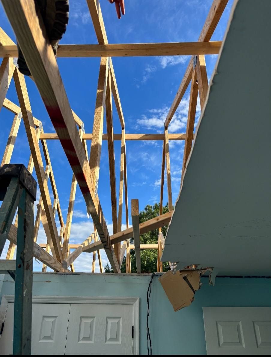 Wooden roof trusses under construction against a blue sky. Interior room visible below.
