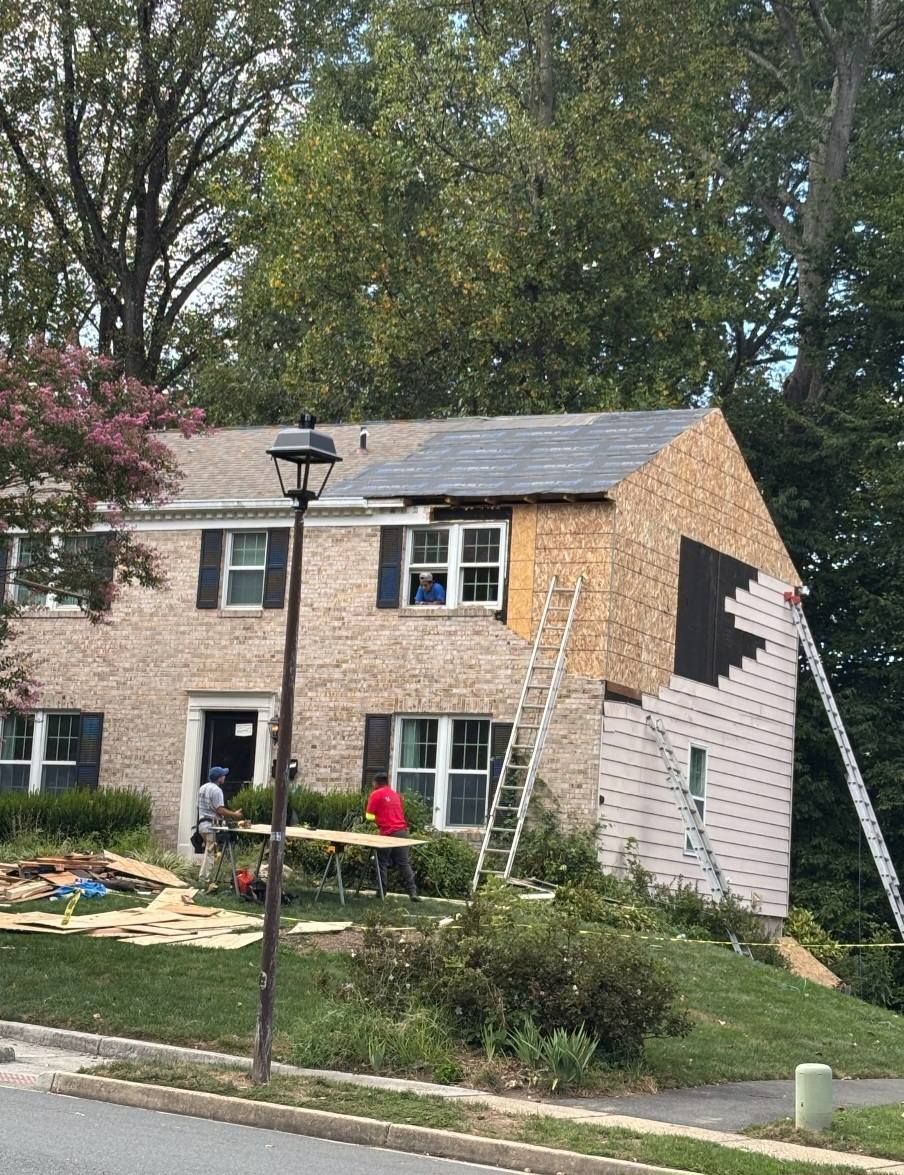 House under construction; workers on ladders, new siding installed. Trees in background.