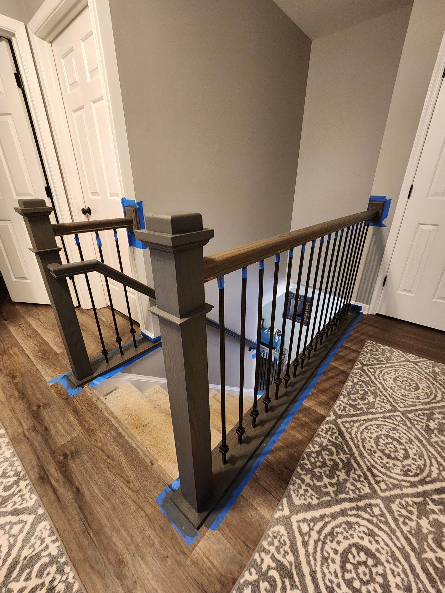Staircase with gray balusters, wood railing, and patterned rugs. Blue tape protects paint during a renovation.