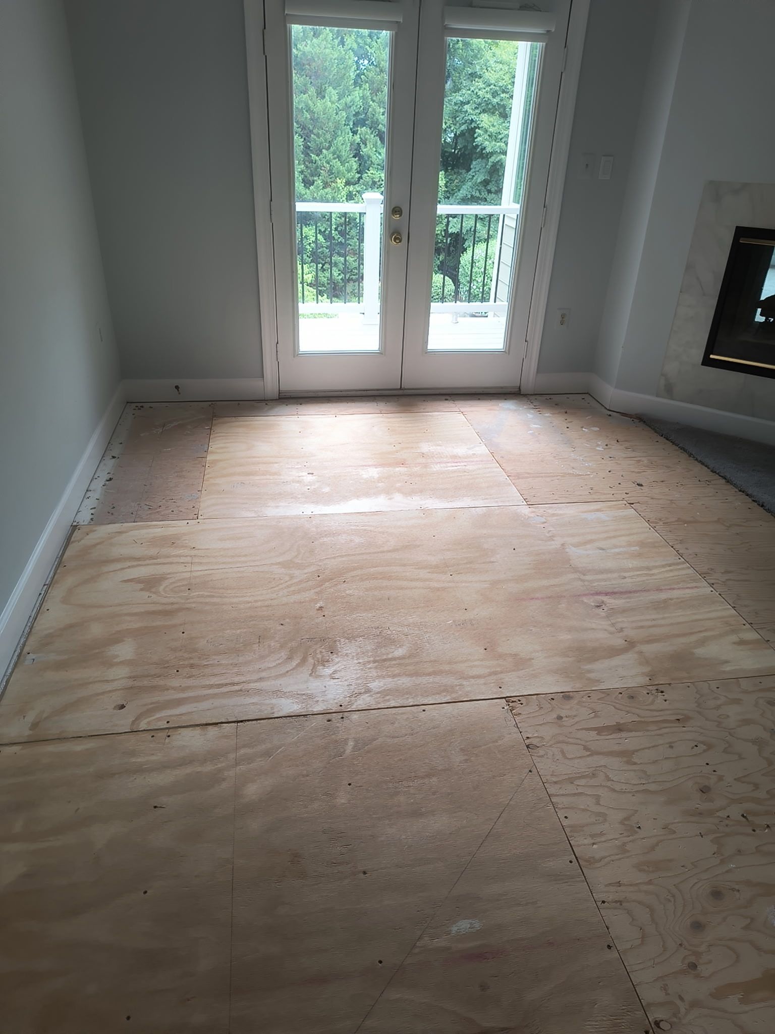 Empty room with bare plywood flooring and French doors opening to a green patio.