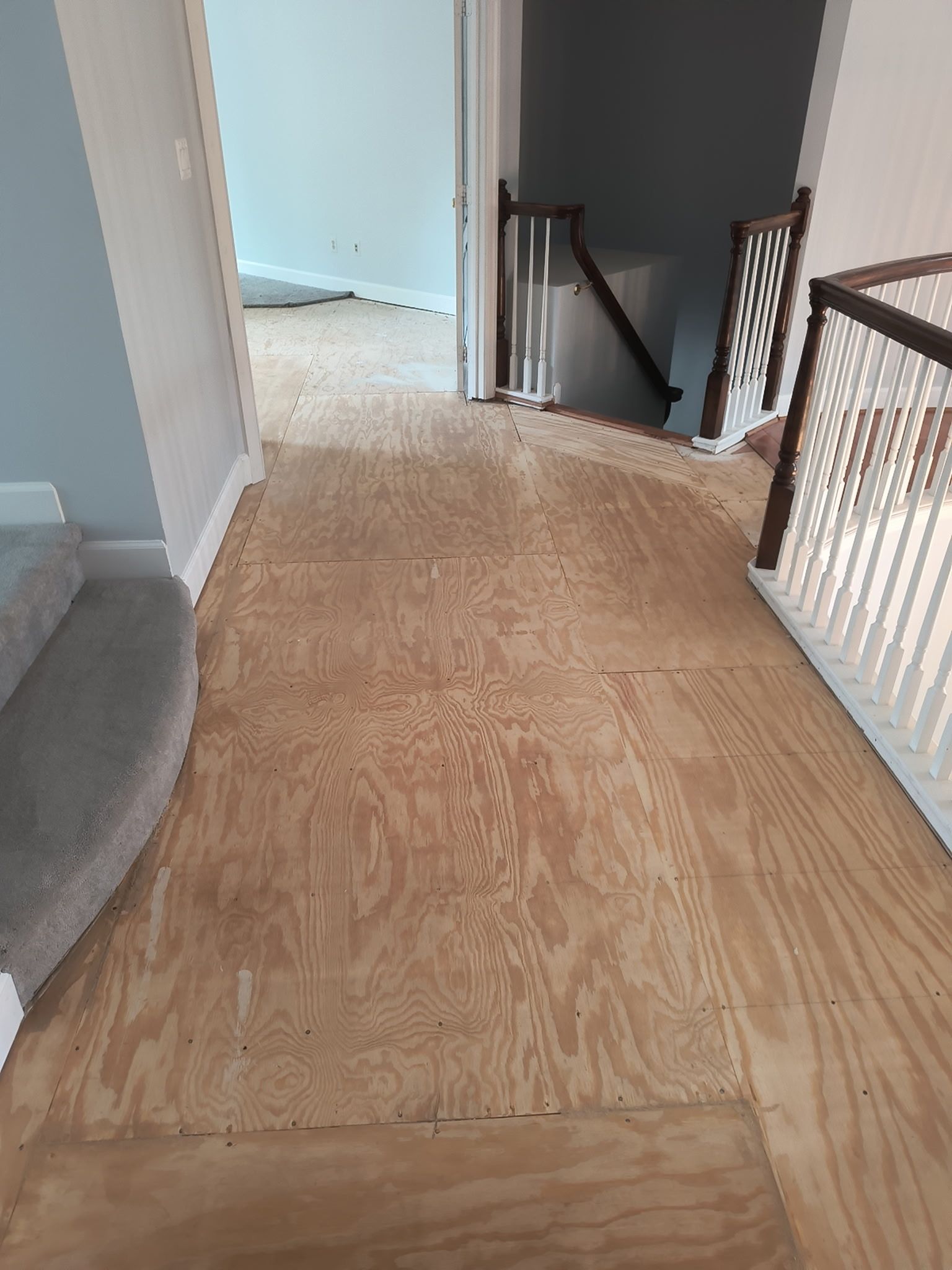 Hallway with plywood floor repair, gray wall, and stair railing leading down.
