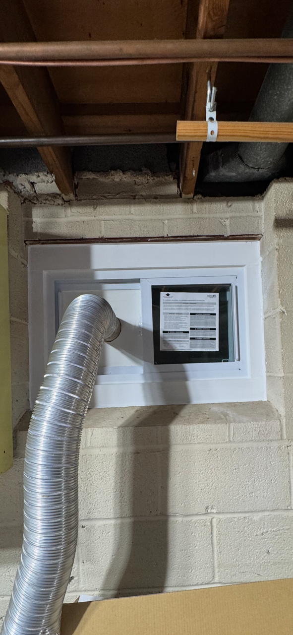 A white window with a black framed sign in a brick basement. A silver exhaust pipe is connected to the window.