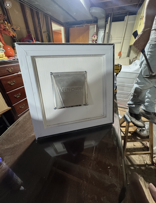 A white-framed ventilation cover is installed on a wall, likely in a basement or utility space. A person is standing nearby holding a hammer.