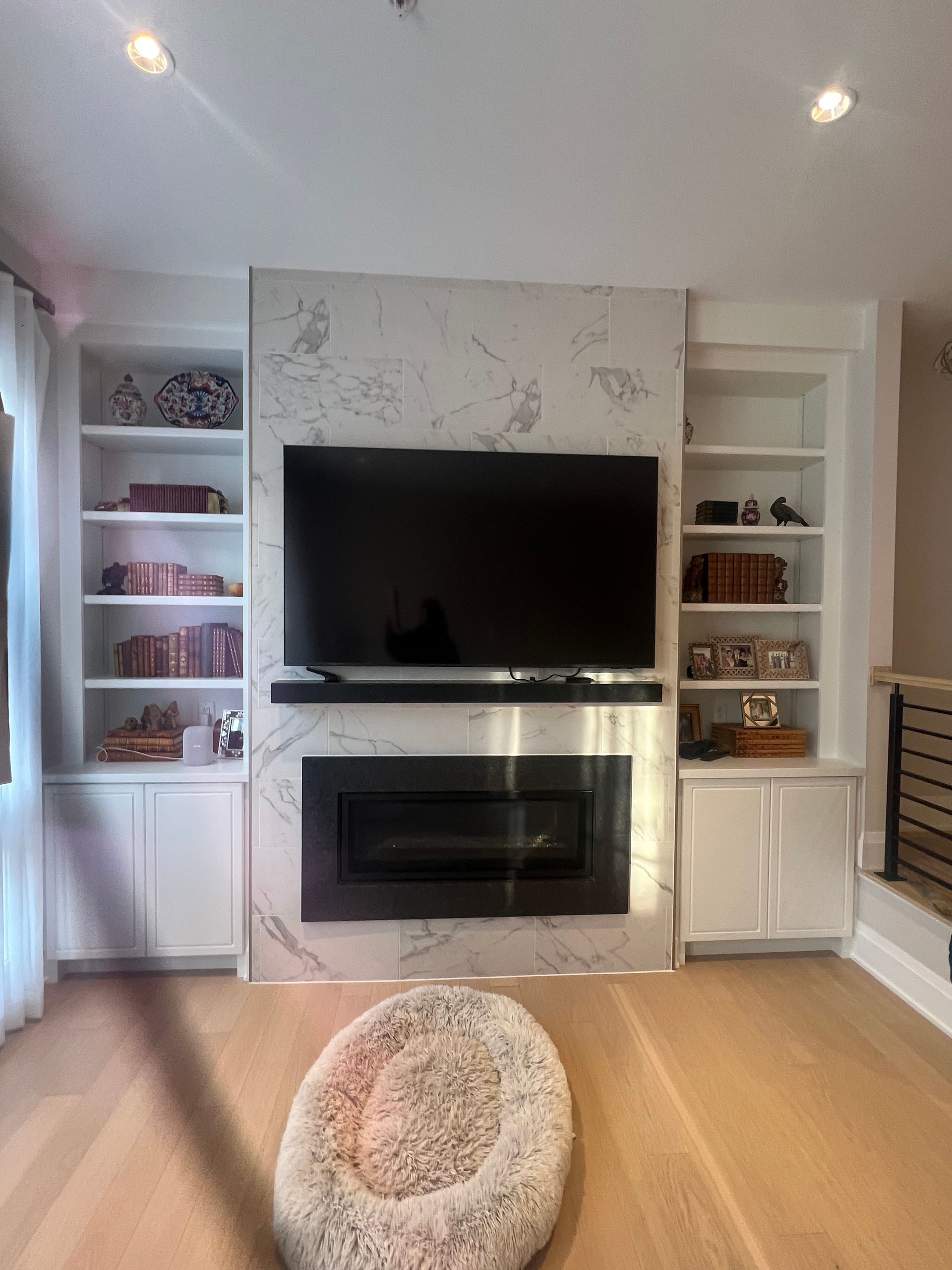 Built-in white shelving and cabinetry frame a fireplace and TV against a marble accent wall. A pet bed is in the foreground.
