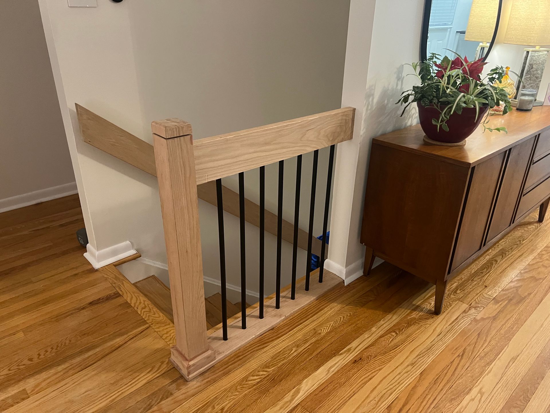 Wooden staircase with black railings. Oak handrail and newel post. Entryway with wood floor, cabinet, and plants.